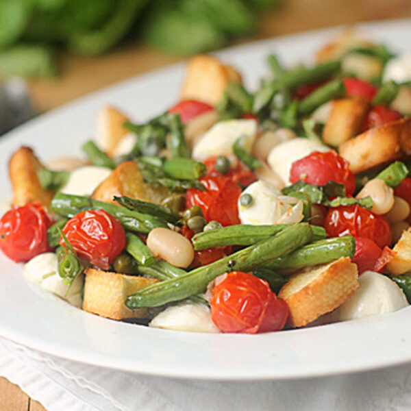 A white plate filled with a salad of roasted cherry tomatoes, green beans, mozzarella balls, white beans, capers, and toasted bread cubes on a light wooden table.