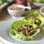 Two lettuce wraps filled with a minced meat and herb mixture are served on a white plate with a wedge of lime. Other dishes and ingredients are blurred in the background.