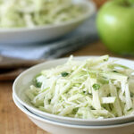 A white bowl filled with shredded cabbage and green apple slaw sits on a wooden surface, with a green apple and another bowl of slaw blurred in the background.