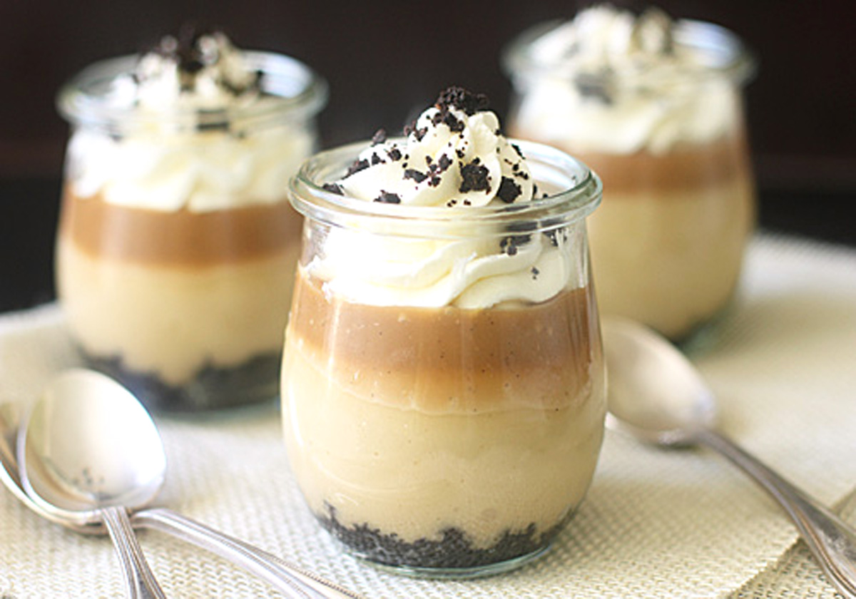Three glass jars filled with layered dessert, including a cookie crumb base, creamy middle, caramel layer, and whipped cream topped with cookie crumbs. Three metal spoons rest nearby on a light textured surface.