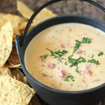 A black pot filled with creamy queso dip topped with chopped cilantro, next to a pile of crispy tortilla chips on a woven placemat.
