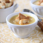 A white bowl filled with creamy soup topped with golden croutons sits on a yellow patterned napkin, with another bowl and two wooden-handled spoons in the background.
