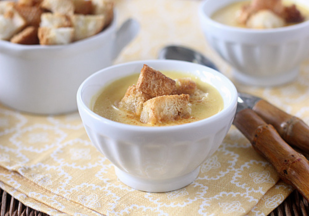 A white bowl filled with creamy soup topped with golden croutons sits on a yellow patterned napkin, with another bowl and two wooden-handled spoons in the background.