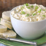 A white bowl filled with a creamy cheese spread topped with chopped green onions sits on a green plate next to several slices of French bread and a butter knife.