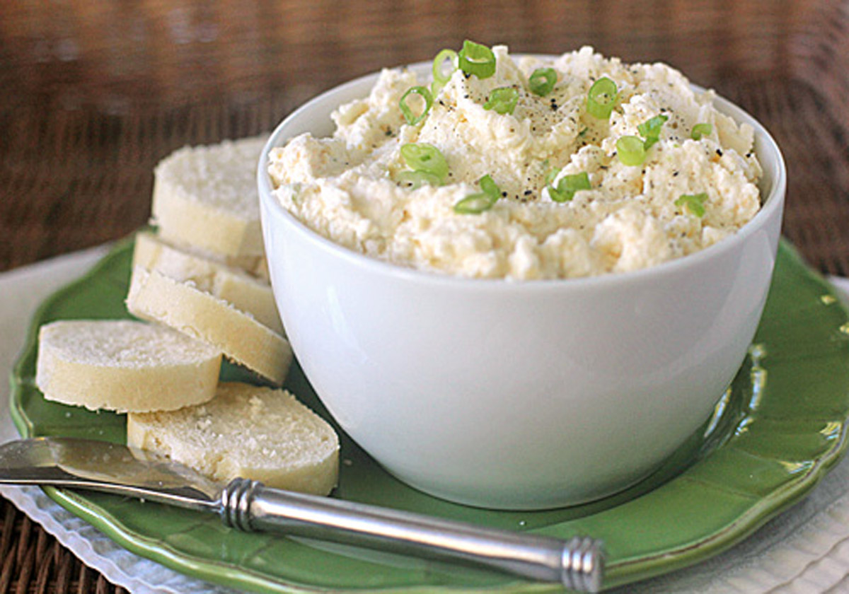A white bowl filled with a creamy cheese spread topped with chopped green onions sits on a green plate next to several slices of French bread and a butter knife.