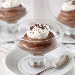 Three glass dessert cups filled with chocolate mousse, each topped with whipped cream and chocolate shavings, placed on white plates with a spoon beside each cup.