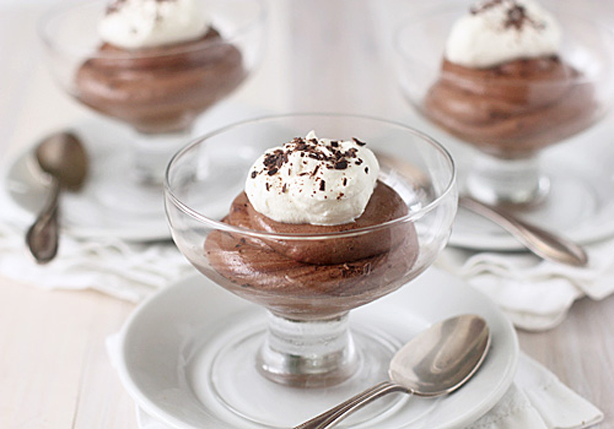 Three glass dessert cups filled with chocolate mousse, each topped with whipped cream and chocolate shavings, placed on white plates with a spoon beside each cup.