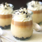 Three glass jars filled with layered dessert featuring a dark crumb base, creamy filling, caramel layer, and whipped cream topped with chocolate crumbs, arranged on a white mat with three spoons nearby.