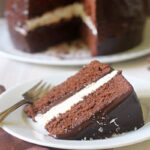 A slice of chocolate cake with white cream filling sits on a plate with a fork. The whole cake, covered in chocolate icing and topped with sprinkles, is in the background with a slice missing.