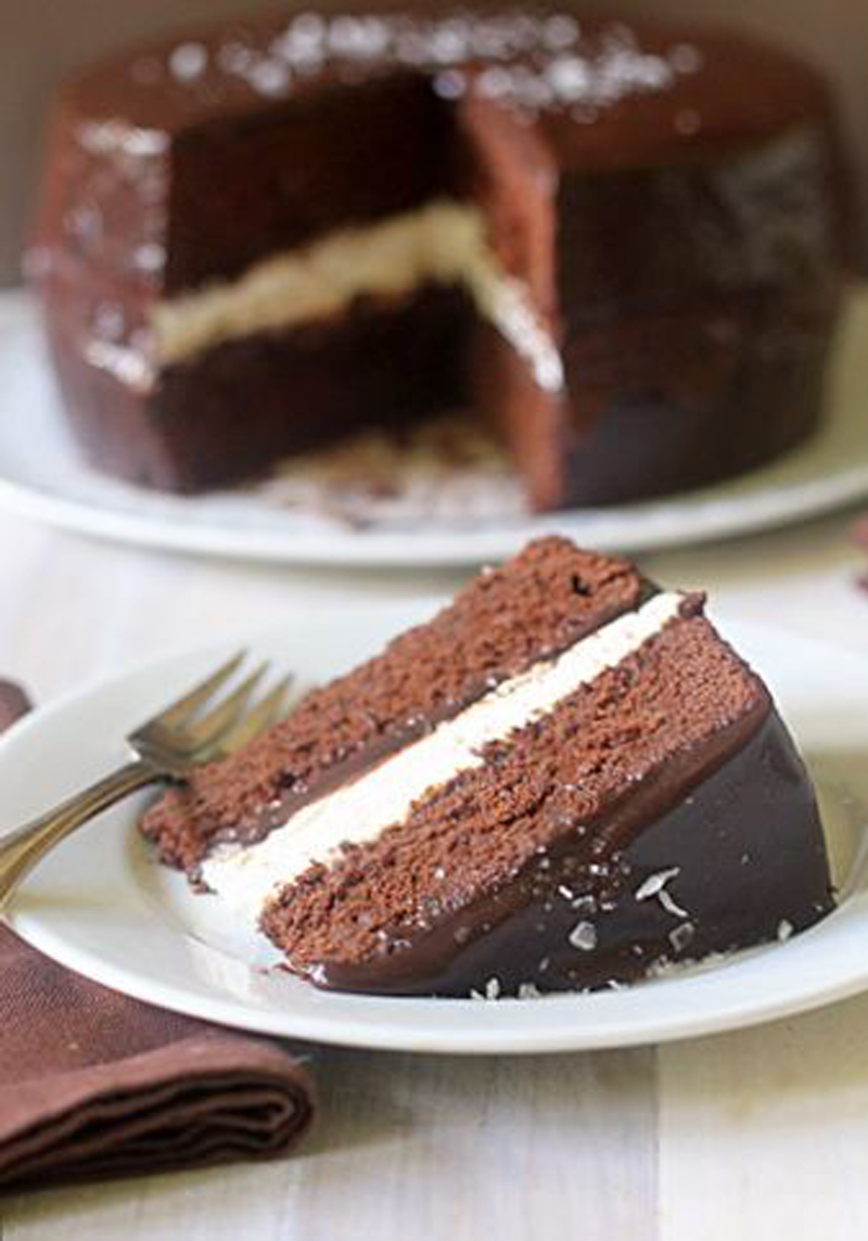 A slice of chocolate cake with white cream filling sits on a plate with a fork. The whole cake, covered in chocolate icing and topped with sprinkles, is in the background with a slice missing.