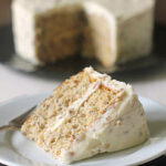 A slice of frosted cake sits on a white plate with a fork, while the rest of the cake, with a missing slice, is in the background. The cake has light-colored layers and white frosting with small specks.
