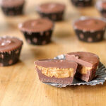 Mini chocolate peanut butter cups on a wooden surface, with one cut in half to show the creamy peanut butter filling inside. Several whole cups are in the background, some in dark liners.