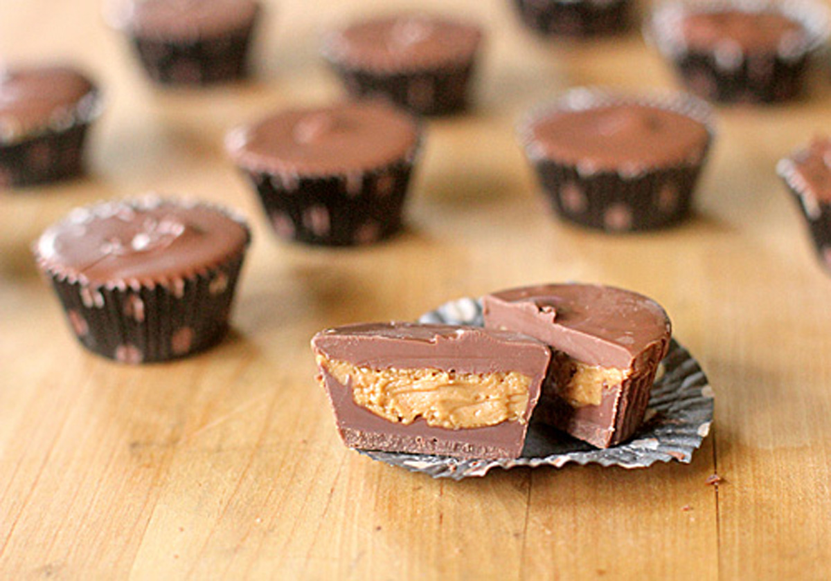 Mini chocolate peanut butter cups on a wooden surface, with one cut in half to show the creamy peanut butter filling inside. Several whole cups are in the background, some in dark liners.