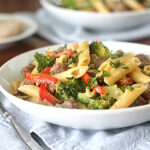 A white bowl filled with penne pasta, broccoli florets, red bell pepper slices, sausage pieces, and fresh herbs sits on a blue napkin next to a fork, with another plate and bread in the background.