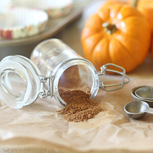 An open glass jar of ground spice spills onto parchment paper beside metal measuring spoons, with small pumpkins and a muffin tin with paper liners in the background.