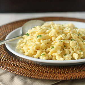 A white plate filled with homemade spaetzle garnished with herbs, placed on a woven placemat with a spoon on the side.