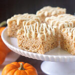 Rice Krispies treats topped with white drizzle are displayed on a white cake stand, with small orange pumpkins arranged around the base, creating an autumn or Halloween-themed setting.