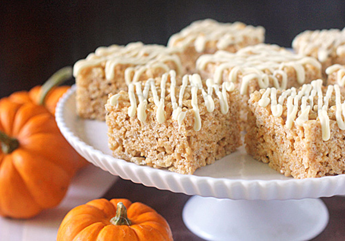 Rice Krispies treats topped with white drizzle are displayed on a white cake stand, with small orange pumpkins arranged around the base, creating an autumn or Halloween-themed setting.