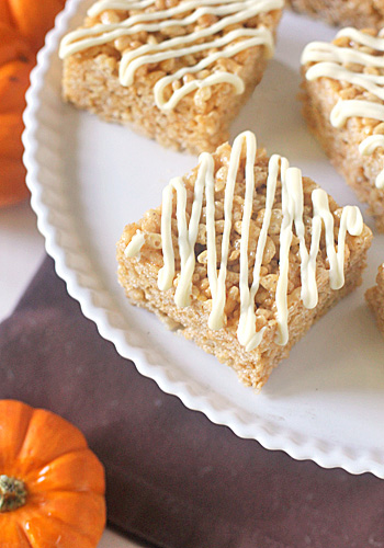 Rice crispy treats topped with white chocolate drizzle arranged on a white plate, with small decorative pumpkins nearby.