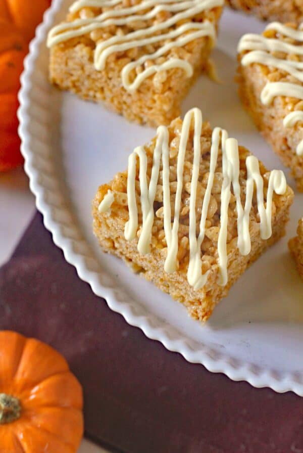 Spiced Pumpkin Rice Krispie Treats, each topped with a delicate white chocolate drizzle, are arranged on a white plate. Small pumpkins are visible nearby, suggesting a festive fall or Halloween theme.