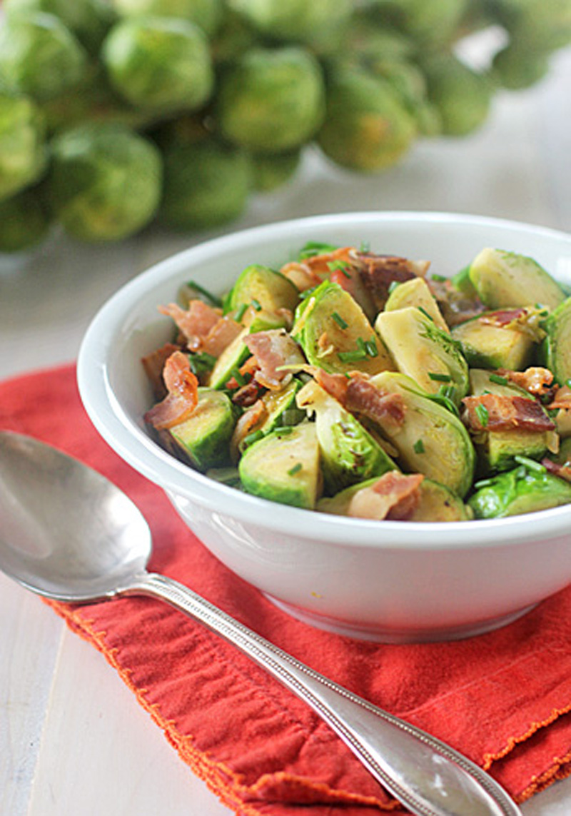 A white bowl filled with sliced Brussels sprouts and pieces of crispy bacon sits on a red cloth napkin, next to a silver spoon. A bundle of fresh Brussels sprouts is blurred in the background.