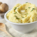 A white bowl filled with creamy mashed potatoes, topped with chopped chives and a small pat of melting butter, sits on a cloth napkin next to a wooden spoon and several whole potatoes in the background.