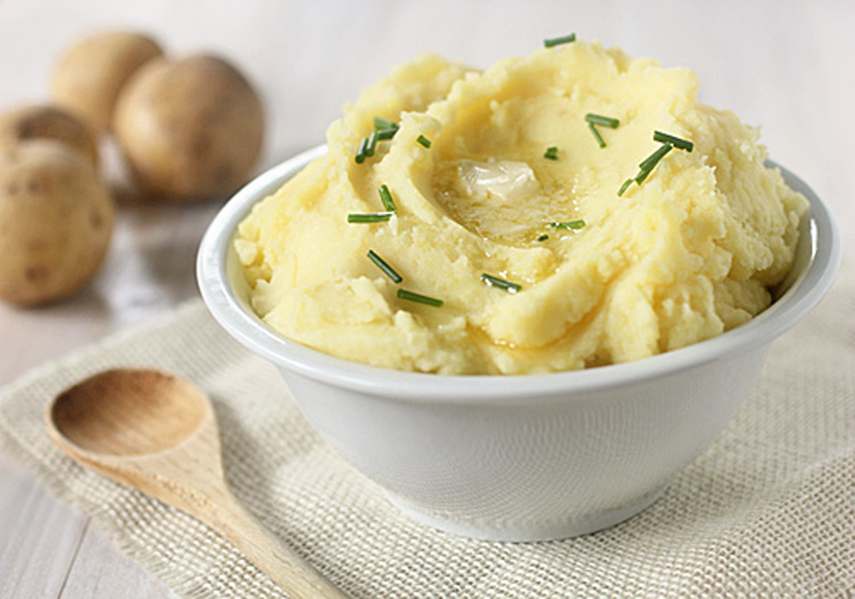 A white bowl filled with creamy mashed potatoes, topped with chopped chives and a small pat of melting butter, sits on a cloth napkin next to a wooden spoon and several whole potatoes in the background.