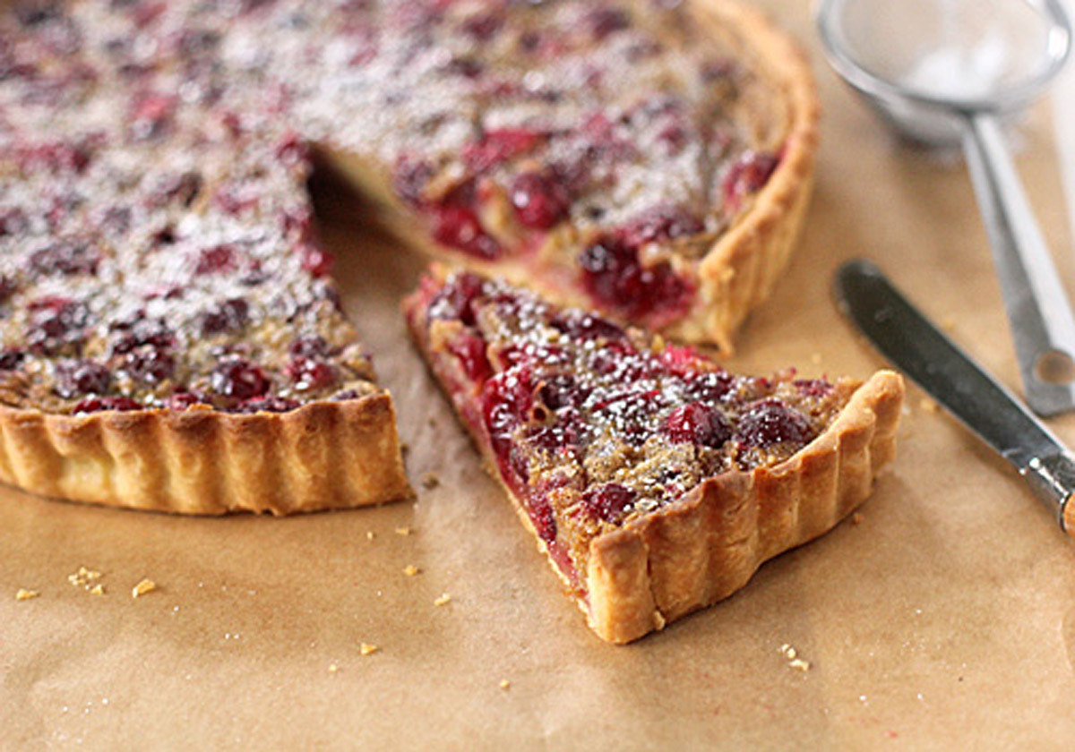 A cranberry tart with a golden crust sits on parchment paper. One slice is cut and slightly separated, showing the berry filling. A knife and a mesh strainer with powdered sugar are nearby.