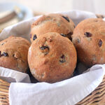A wicker basket lined with a white cloth holds four round, golden-brown raisin bread rolls with visible raisins baked into the surface. The background is softly blurred with tableware visible.