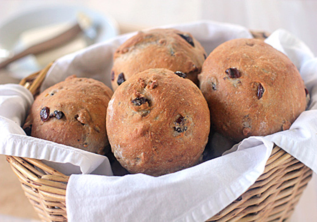 A wicker basket lined with a white cloth holds four round, golden-brown raisin bread rolls with visible raisins baked into the surface. The background is softly blurred with tableware visible.