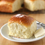 A soft, fluffy dinner roll with a golden-brown top is served on a white plate next to a pat of butter, with more rolls in the background on a wooden surface.