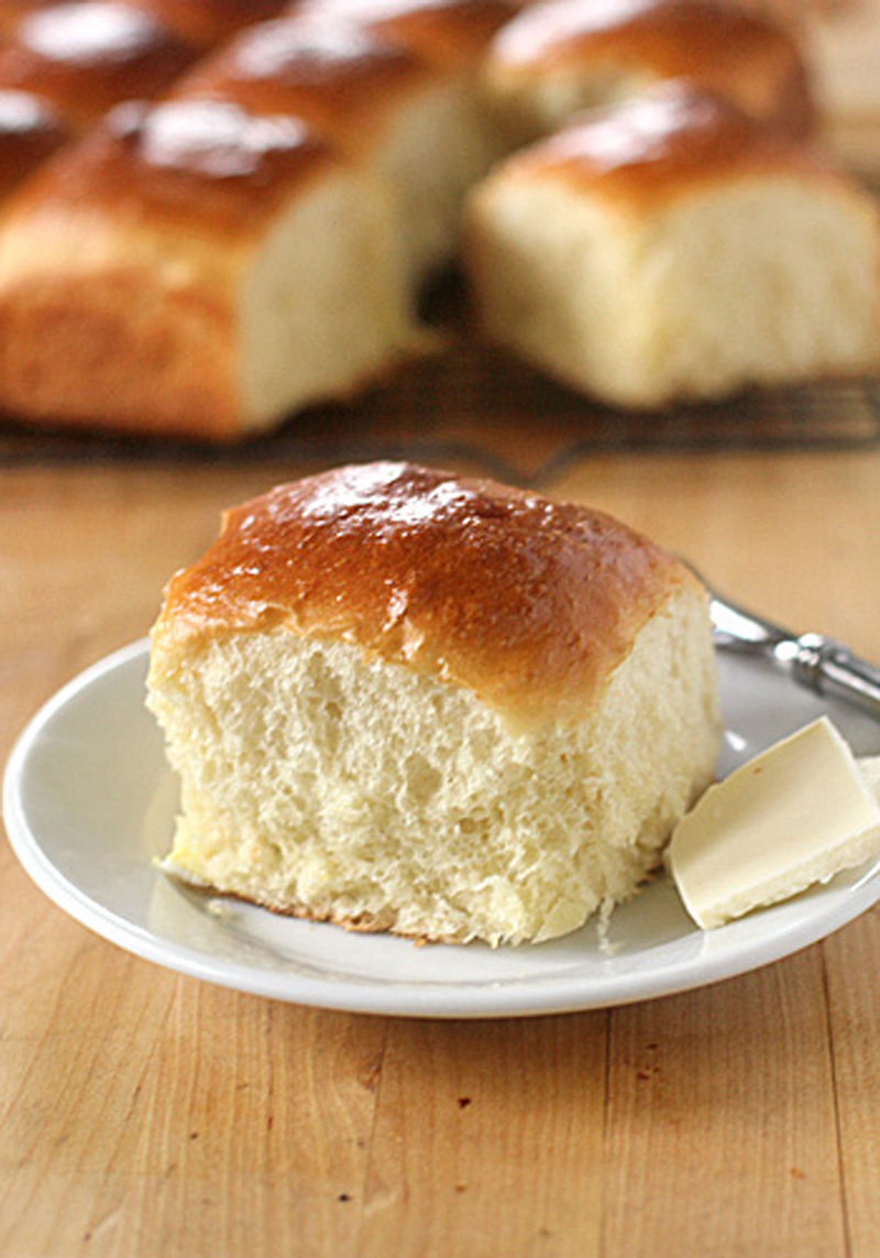 A soft, fluffy dinner roll with a golden-brown top is served on a white plate next to a pat of butter, with more rolls in the background on a wooden surface.