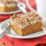 A slice of pumpkin cake topped with cream cheese and toasted coconut sits on a white plate with a silver fork, placed on a red napkin. Another plate with cake and a white mug are visible in the background.