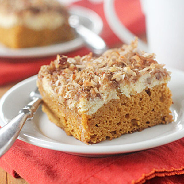 A slice of pumpkin cake topped with cream cheese and toasted coconut sits on a white plate with a silver fork, placed on a red napkin. Another plate with cake and a white mug are visible in the background.