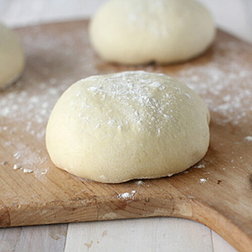 Three balls of dough dusted with flour rest on a wooden surface, ready for baking. The background is softly blurred, highlighting the smooth texture of the dough in the foreground.