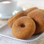 A white plate with several sugar-coated donuts sits on a folded napkin. In the background, there is a white cup and saucer filled with a hot beverage, all set on a light, patterned tablecloth.