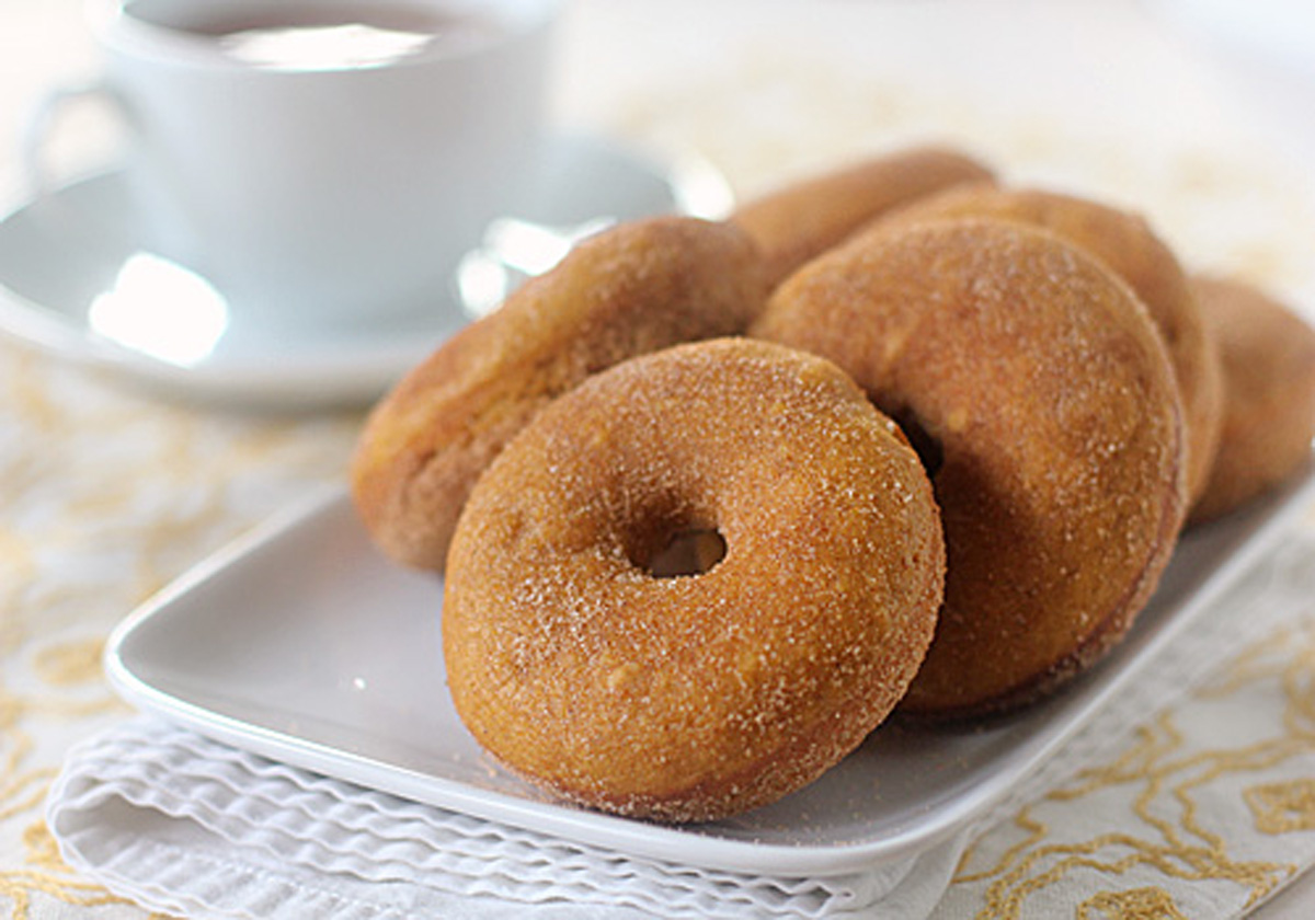 A white plate with several sugar-coated donuts sits on a folded napkin. In the background, there is a white cup and saucer filled with a hot beverage, all set on a light, patterned tablecloth.