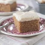 A slice of frosted spice cake sits on a decorative plate with a fork, atop a folded cloth napkin. Another piece of cake and a baking pan are blurred in the background.