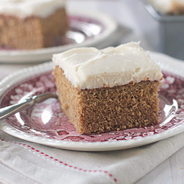 A slice of frosted spice cake sits on a decorative plate with a fork, atop a folded cloth napkin. Another piece of cake and a baking pan are blurred in the background.