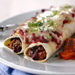Two enchiladas filled with beans and meat, topped with melted cheese, tomato sauce, and chopped green onions, served on a white plate with a side of roasted vegetables.