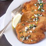 Two breaded and fried cutlets garnished with capers and herbs are served on a white plate with a lemon wedge and a serving fork, set on a dark textured tablecloth.