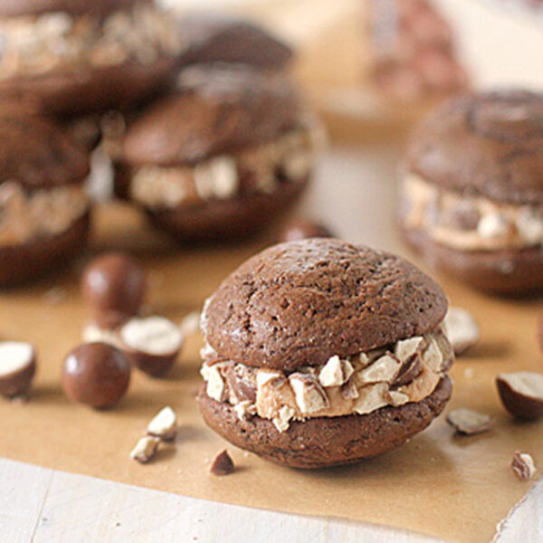 Five chocolate whoopie pies with cream filling and chopped chocolate pieces, arranged on a sheet of parchment paper with a few scattered chocolate-coated candies around them.