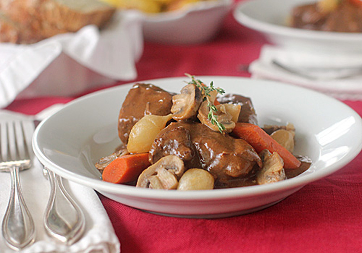 A white bowl filled with beef stew, carrots, mushrooms, and pearl onions, garnished with fresh thyme, sits on a red tablecloth next to silverware and a white napkin.