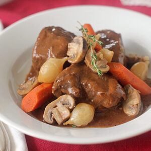 A white bowl filled with beef stew, featuring chunks of beef, carrots, mushrooms, pearl onions, and a rich brown sauce, garnished with a sprig of fresh thyme.
