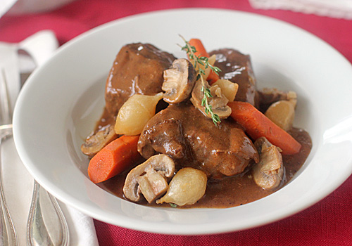 A white bowl filled with beef stew, featuring chunks of beef, carrots, mushrooms, pearl onions, and a rich brown sauce, garnished with a sprig of fresh thyme.
