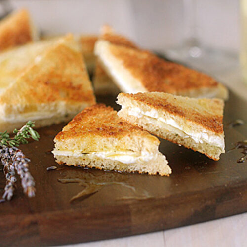 Triangular grilled cheese sandwiches are arranged on a wooden board, with a sprig of herbs and a drizzle of honey nearby. A glass of white wine is blurred in the background.