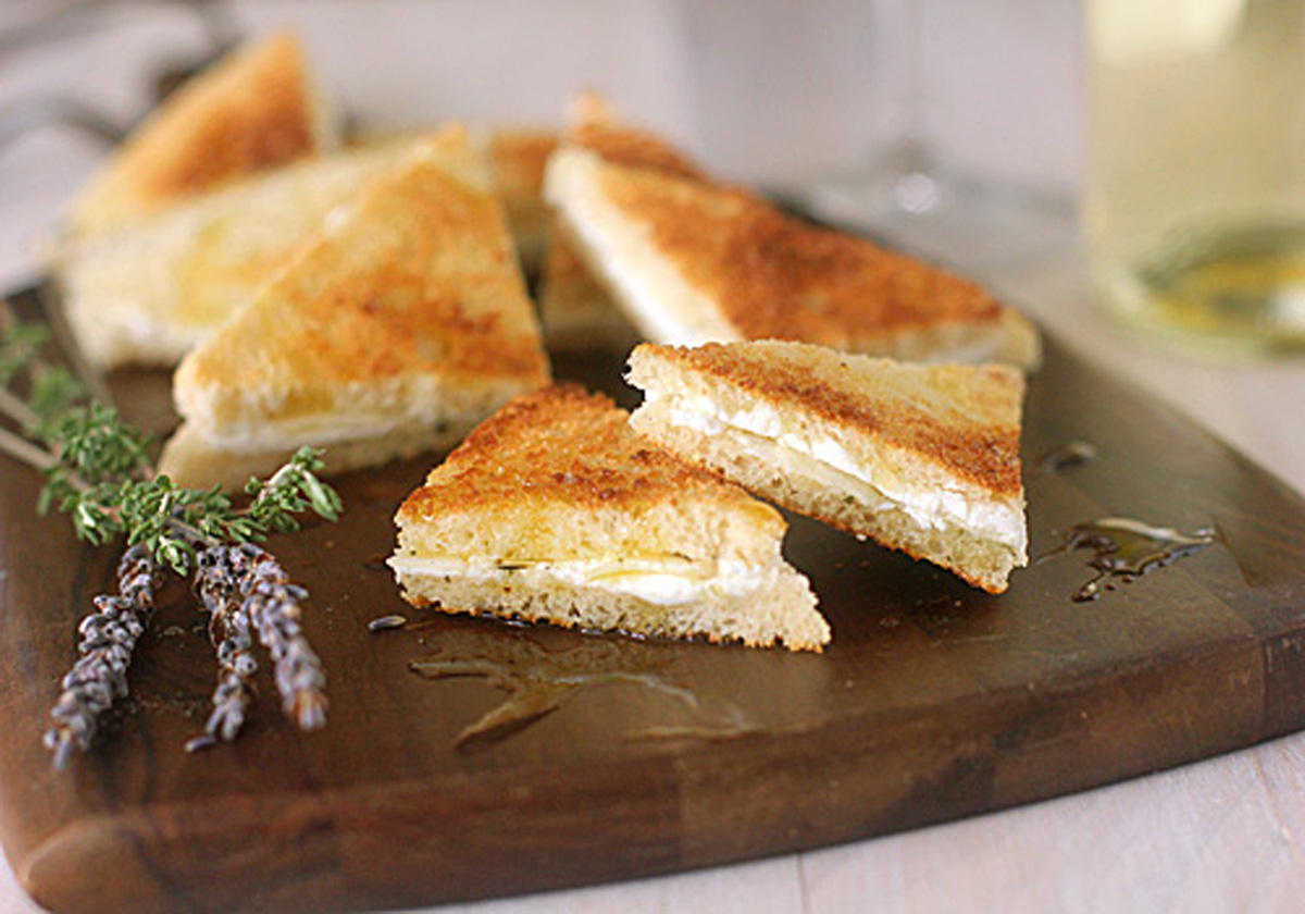 Triangular grilled cheese sandwiches are arranged on a wooden board, with a sprig of herbs and a drizzle of honey nearby. A glass of white wine is blurred in the background.