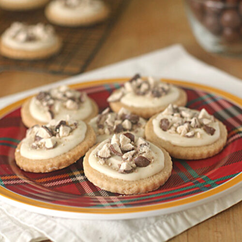 A red plaid plate with six frosted cookies topped with chopped chocolate pieces sits on a white napkin. More cookies and a glass bowl of chocolates are in the blurred background.