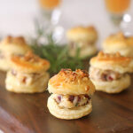 Small, round puff pastry bites filled with a creamy mixture and topped with herbs, arranged on a wooden serving board, with blurred drinks in the background.