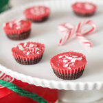 Red and brown chocolate peppermint cups topped with crushed candy canes are arranged on a white cake stand, with two small candy canes crossed in the background.